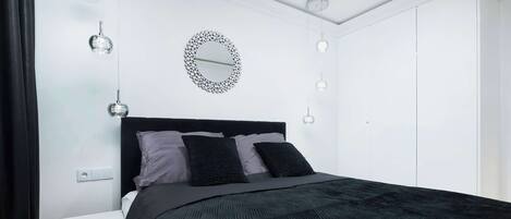 A bedroom with a black bedspread, a round mirror on the wall, and pendant lights.