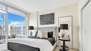 Moderni hotellihuone, jossa on suuri sänky, yöpöytä lampun kanssa, peili ja suuret ikkunat, joista avautuu näkymä kaupunkiin.