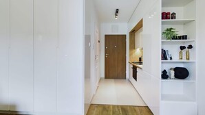 A modern, minimalist hallway with white cabinetry, a wooden door, and a kitchen area with white shelving. - Stylish Studio with Balcony | Modern Building (Warszawa)