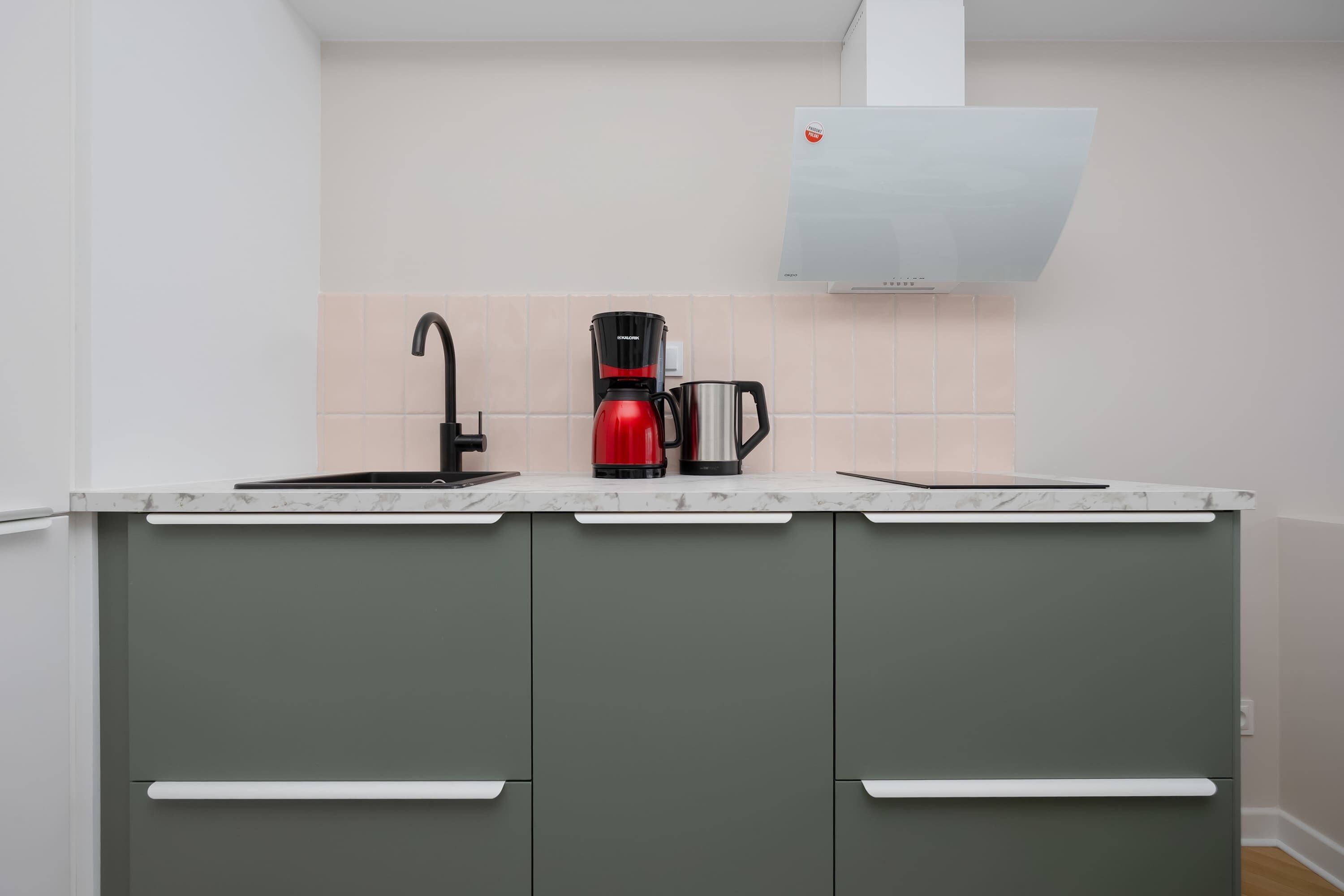 A kitchen with a modern design, featuring a grey cabinet with white handles, a red coffee maker, and a stainless steel coffee pot.