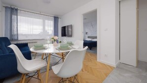 Un salon moderne avec un coin repas, comprenant un canapé bleu, des chaises blanches, une table ronde avec des assiettes vertes et un téléviseur fixé au mur.