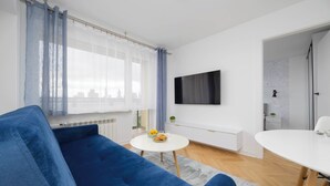 Un salon moderne avec un canapé bleu, une table basse ronde et blanche ornée de fruits, un téléviseur blanc fixé au mur et une table à manger blanche.