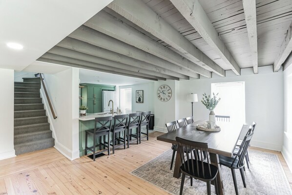 A spacious living area with a wooden staircase, a dining table with chairs, a kitchen with green cabinets, and a large clock on the wall.