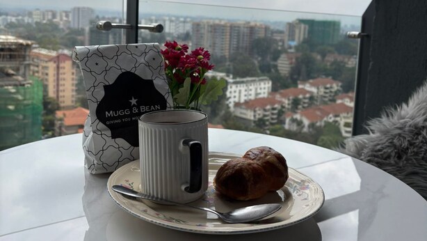 A table with a mug, a pastry, and a bag with a logo, set against a cityscape view. - Spacious,New Apartment with WiFi,Gym and heated pool in Lavington, Nairobi (Nairobi)