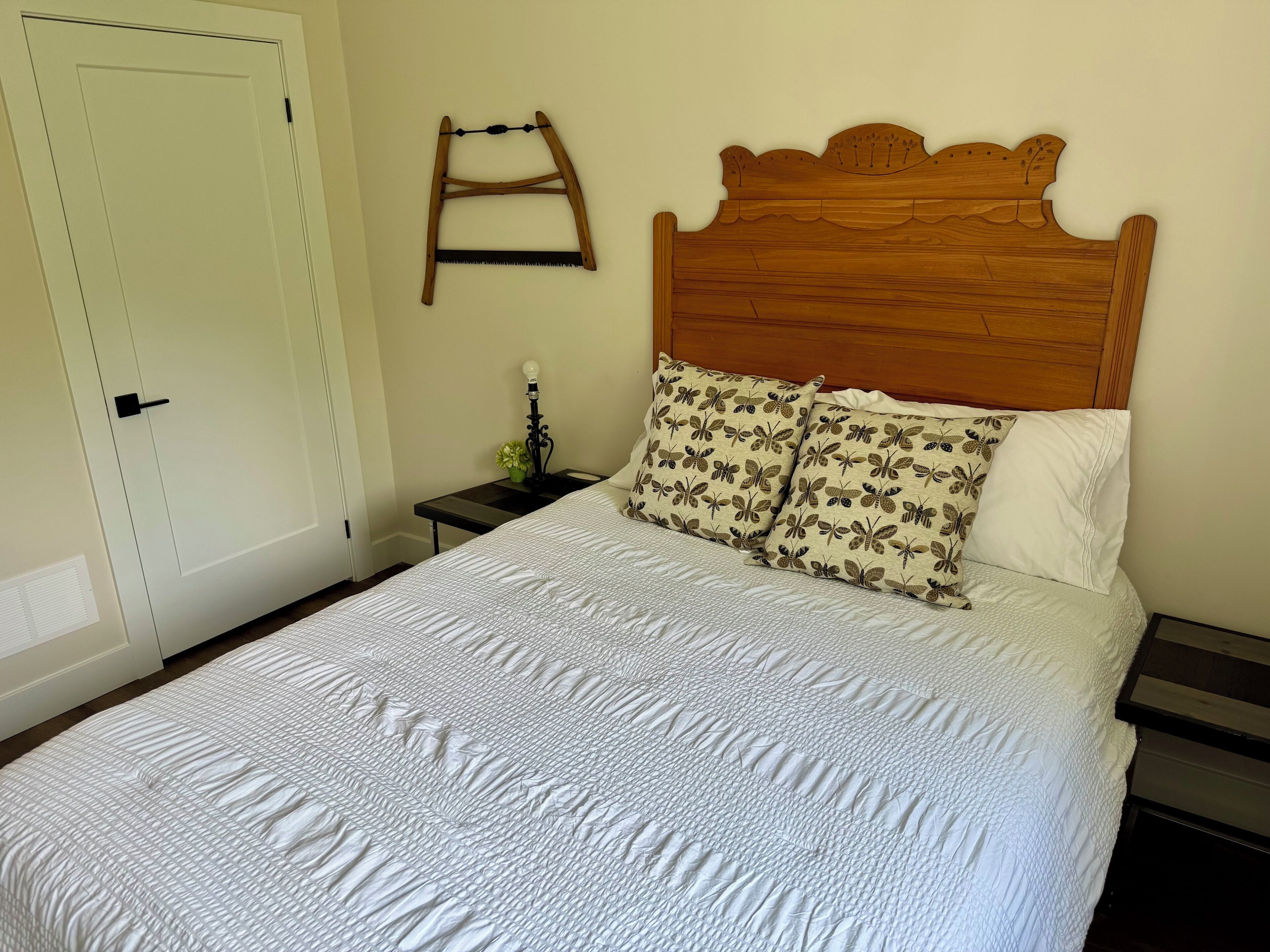 A bedroom with a wooden headboard, a white quilted bedspread, two patterned pillows, a bedside table with a lamp, and a door with a handle.