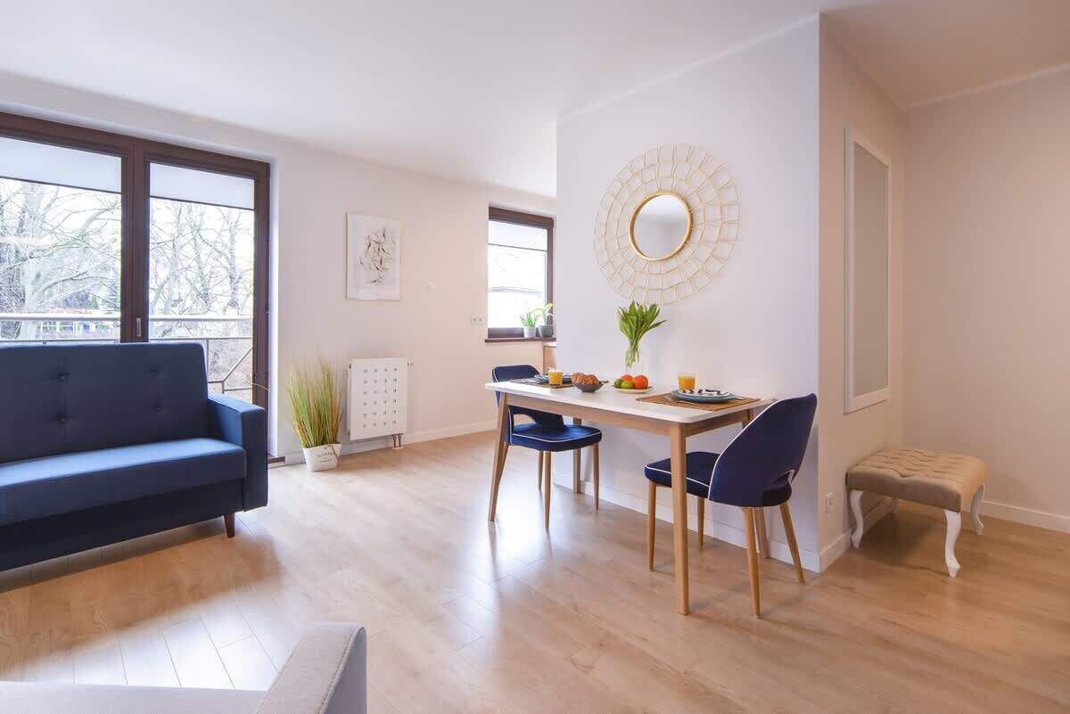 A modern living room with a blue sofa, a dining table with chairs, and a small stool. There is a large window providing natural light and a view of trees outside.