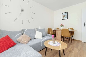 Uma sala de estar moderna com um sofá cinzento, um travesseiro vermelho, uma mesa de jantar de madeira e um relógio de parede.