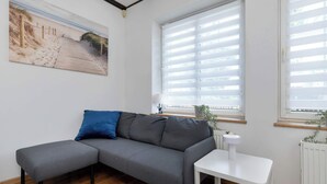 A modern living room with a grey sectional sofa, a small white side table, and a large window with blinds.