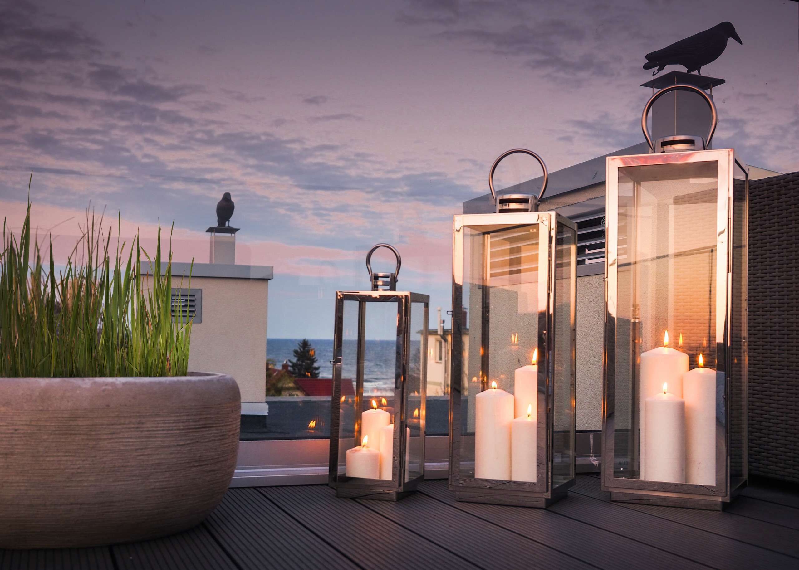 A rooftop terrace with three glass lanterns, each holding a candle, and a large potted plant.