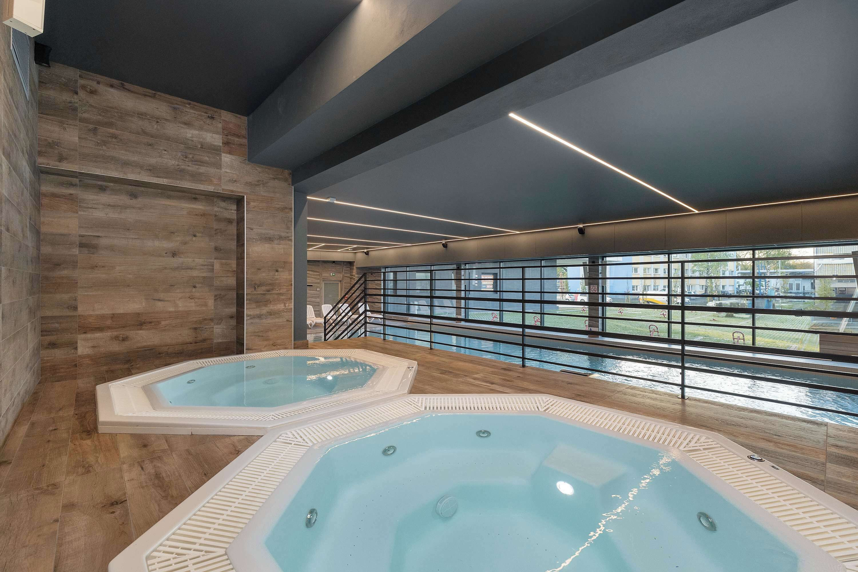 Un espace piscine intérieur moderne, doté de deux bains à remous, d’un plancher en bois et offrant une vue à travers une rambarde en verre.