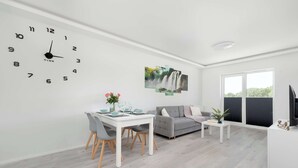 A modern living room with a dining area, a grey sofa, a white coffee table, and a large wall clock.