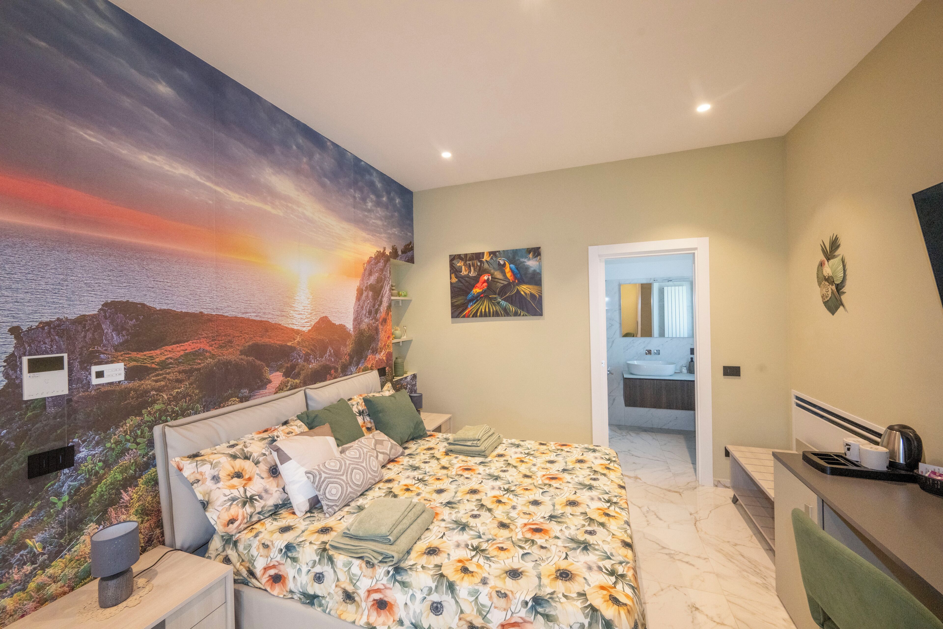 Een slaapkamer met een grote muurschildering van een zonsondergang, een bed met bloemenbeddengoed en een badkamer die door een open deur zichtbaar is.