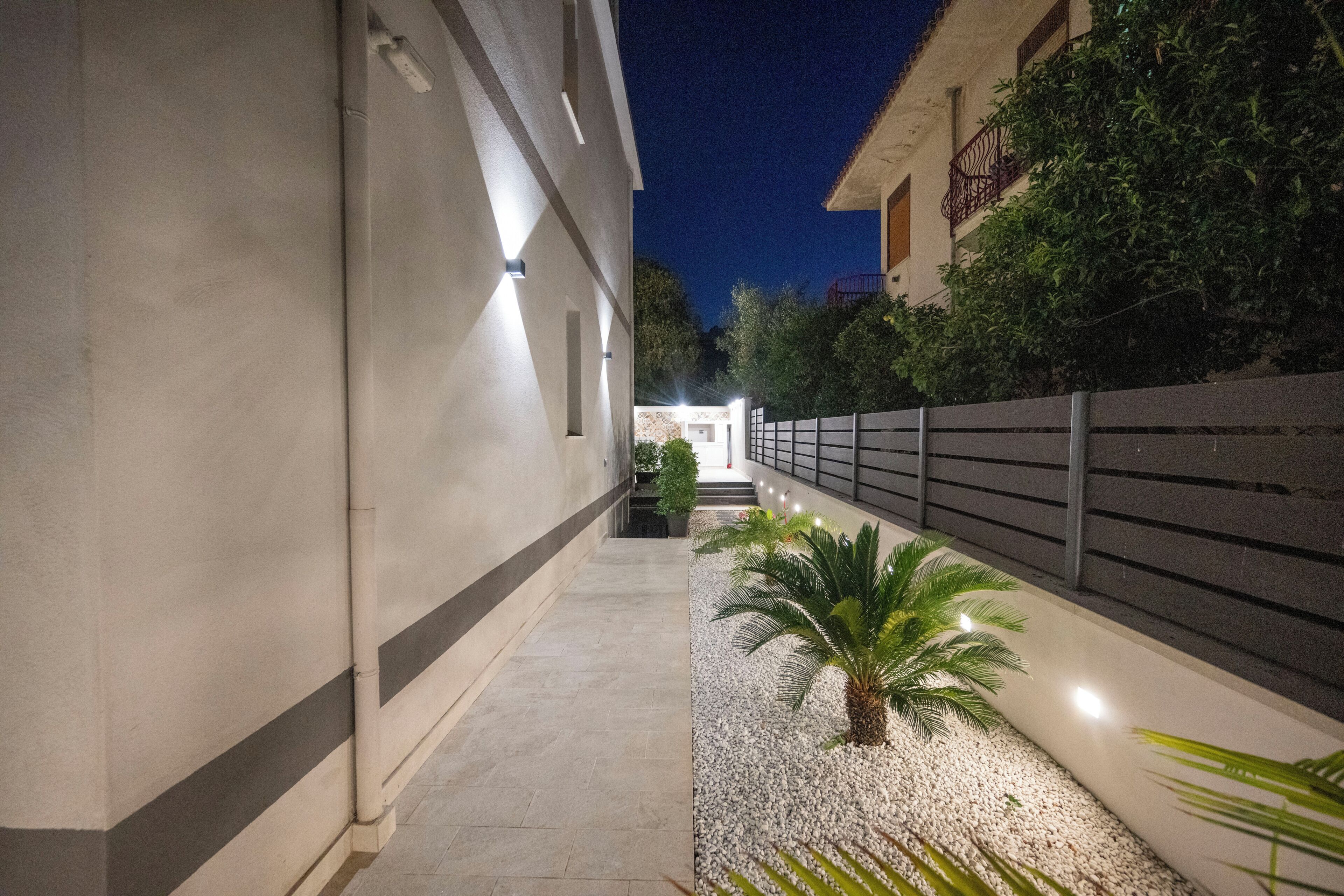 Ein beleuchteter Korridor bei Nacht mit einem Weg, einer Palme und einem Gebäude mit Balkon.