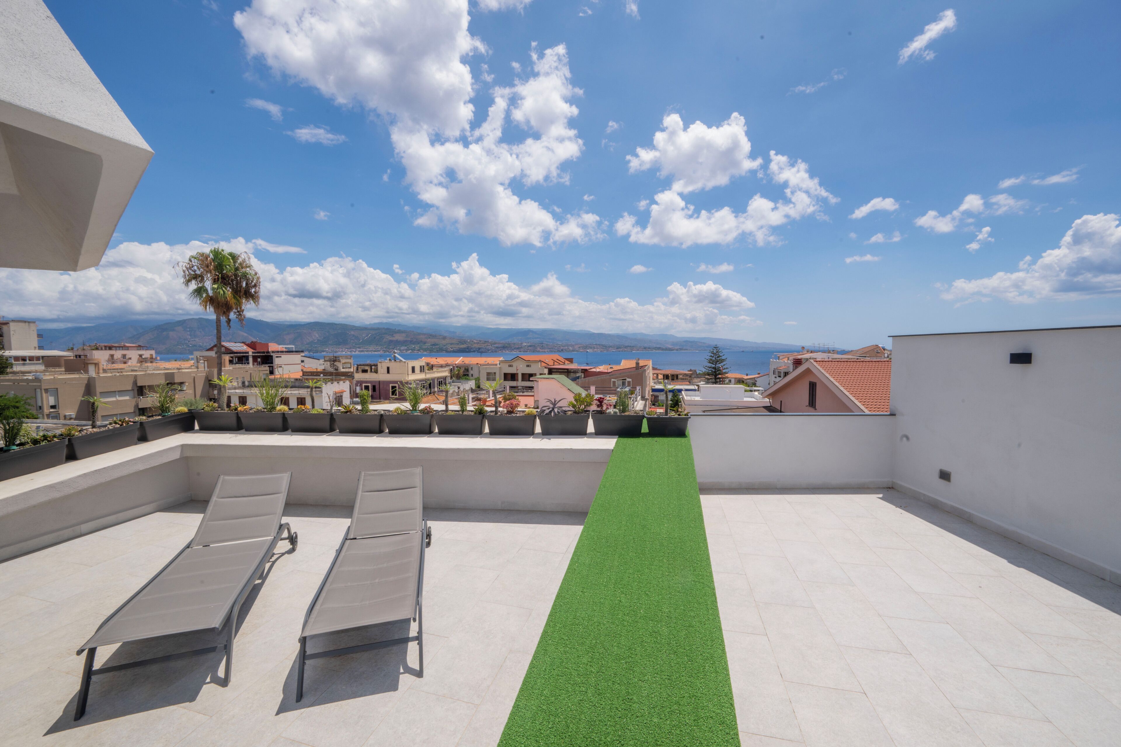 Eine Dachterrasse mit zwei Liegestühlen, einem begrünten Rasenbereich und Blick auf eine Küstenstadt.