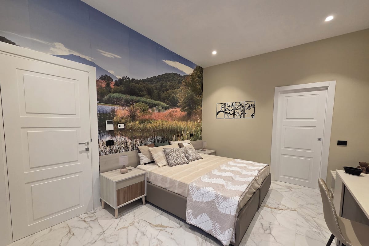 Un dormitorio con cama, mesitas de noche, un escritorio y un mural de un paisaje.