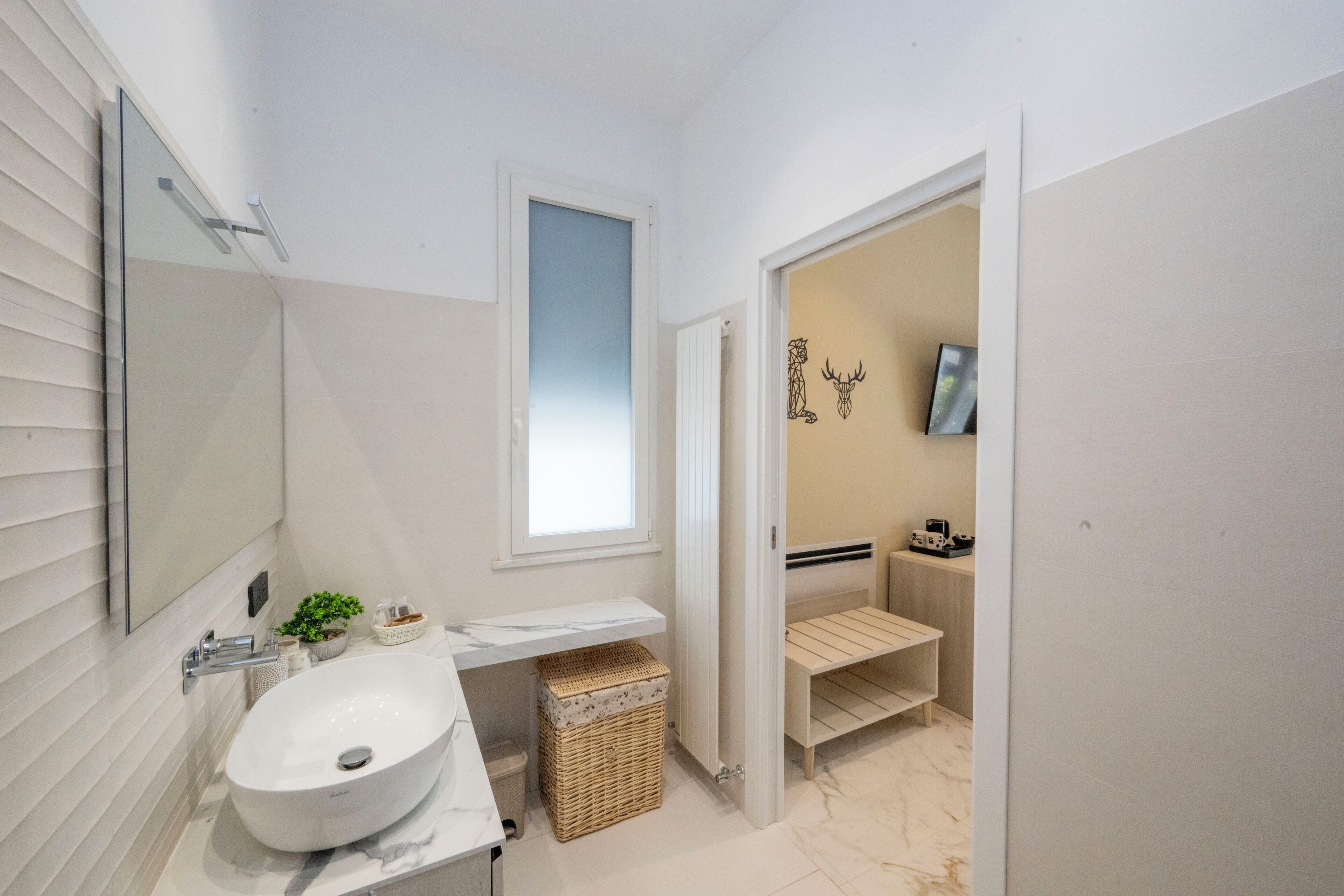 Un baño moderno con un lavamanos blanco grande, un espejo, una ventana y una puerta que da a otra habitación.