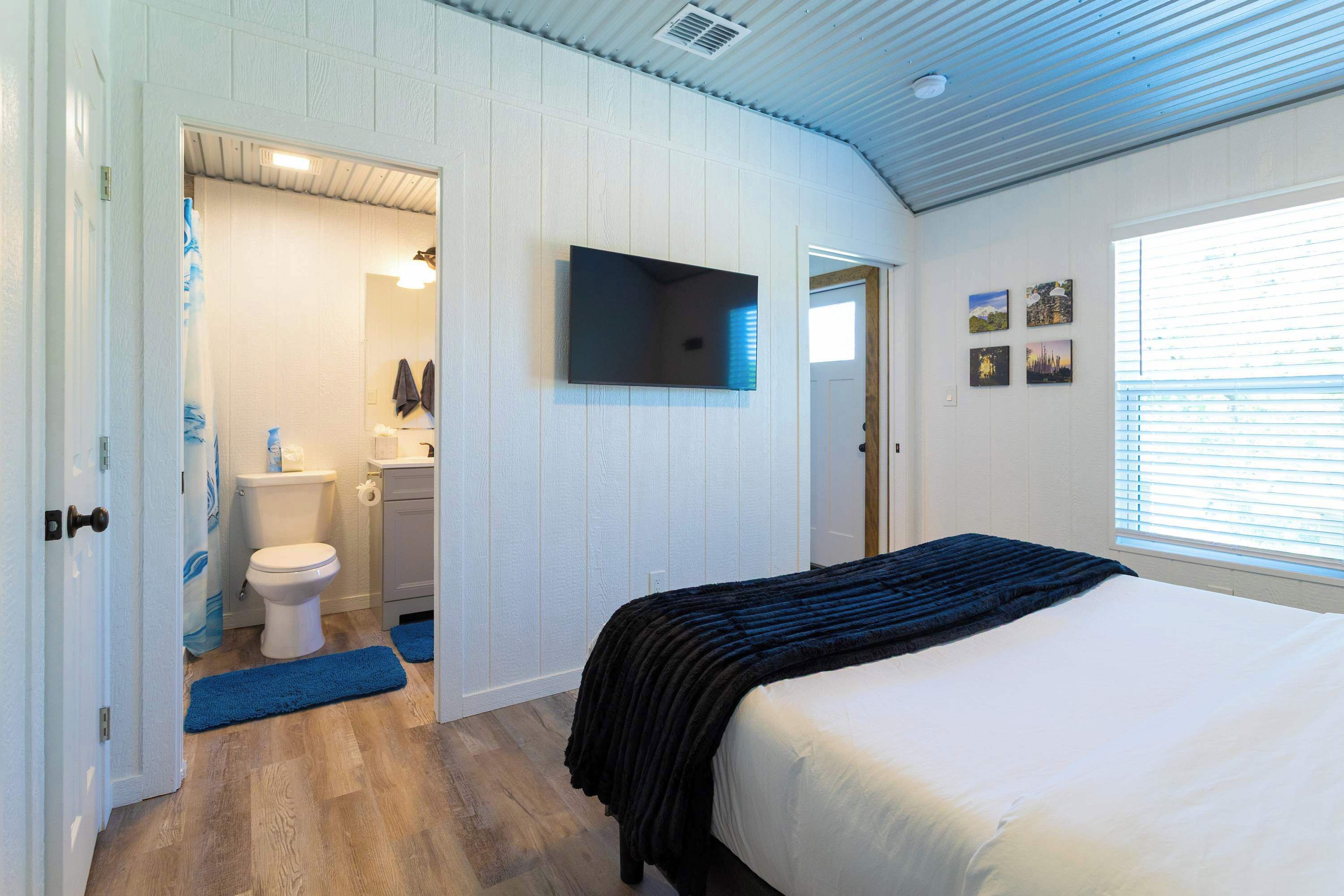 A bedroom with a bed, a TV mounted on the wall, a window with blinds, and a door leading to a bathroom.