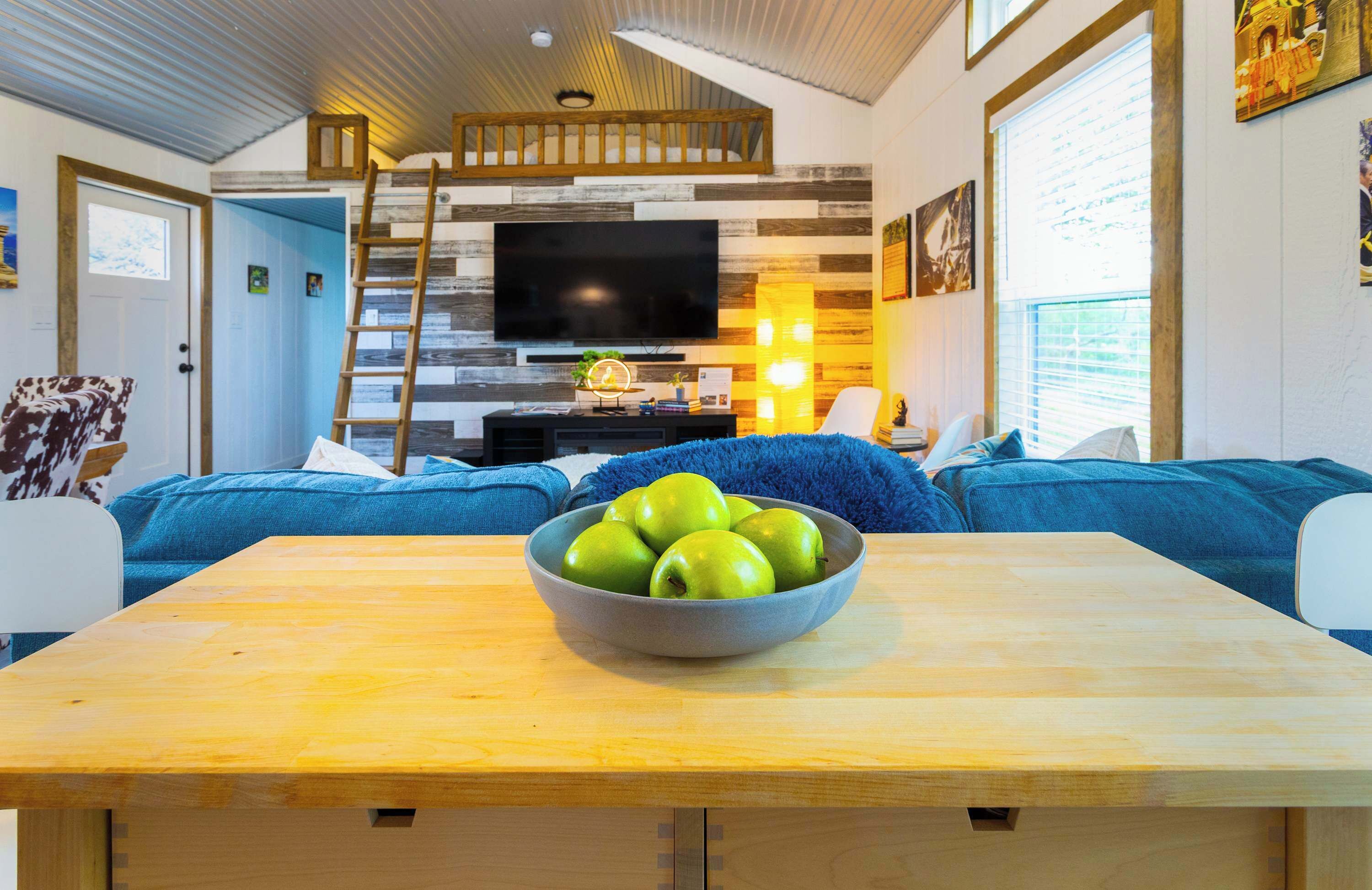 A cozy living room with a wooden table, a bowl of green apples, a TV, and blue sofas.