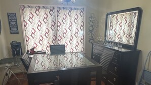 Un comedor con mesa, sillas y una lámpara de araña. Hay un espejo en la pared y un aparador con cajones.