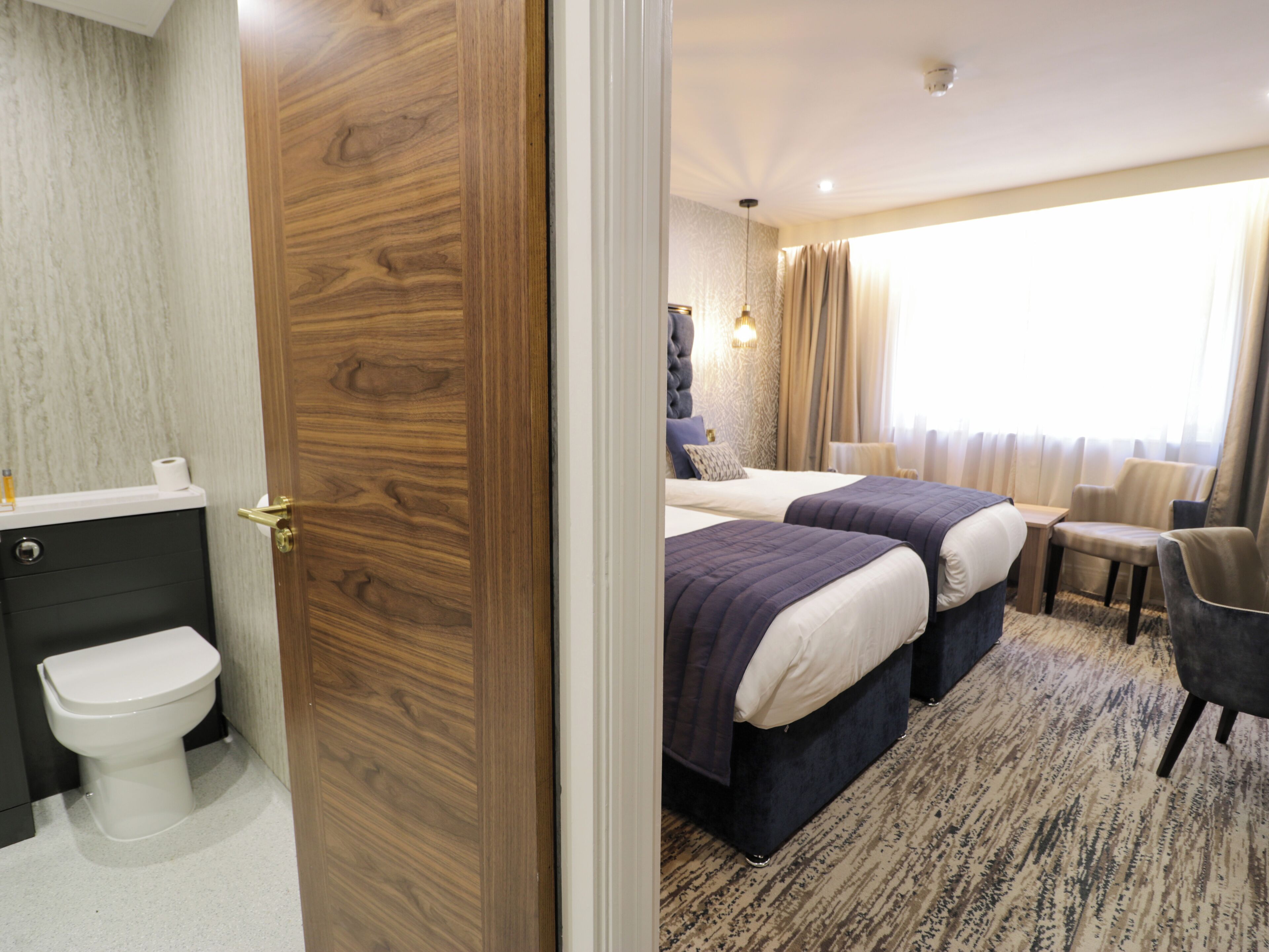 A hotel room with a wooden door, a bathroom with a toilet, two beds with dark bedding, a small table, and a chair.