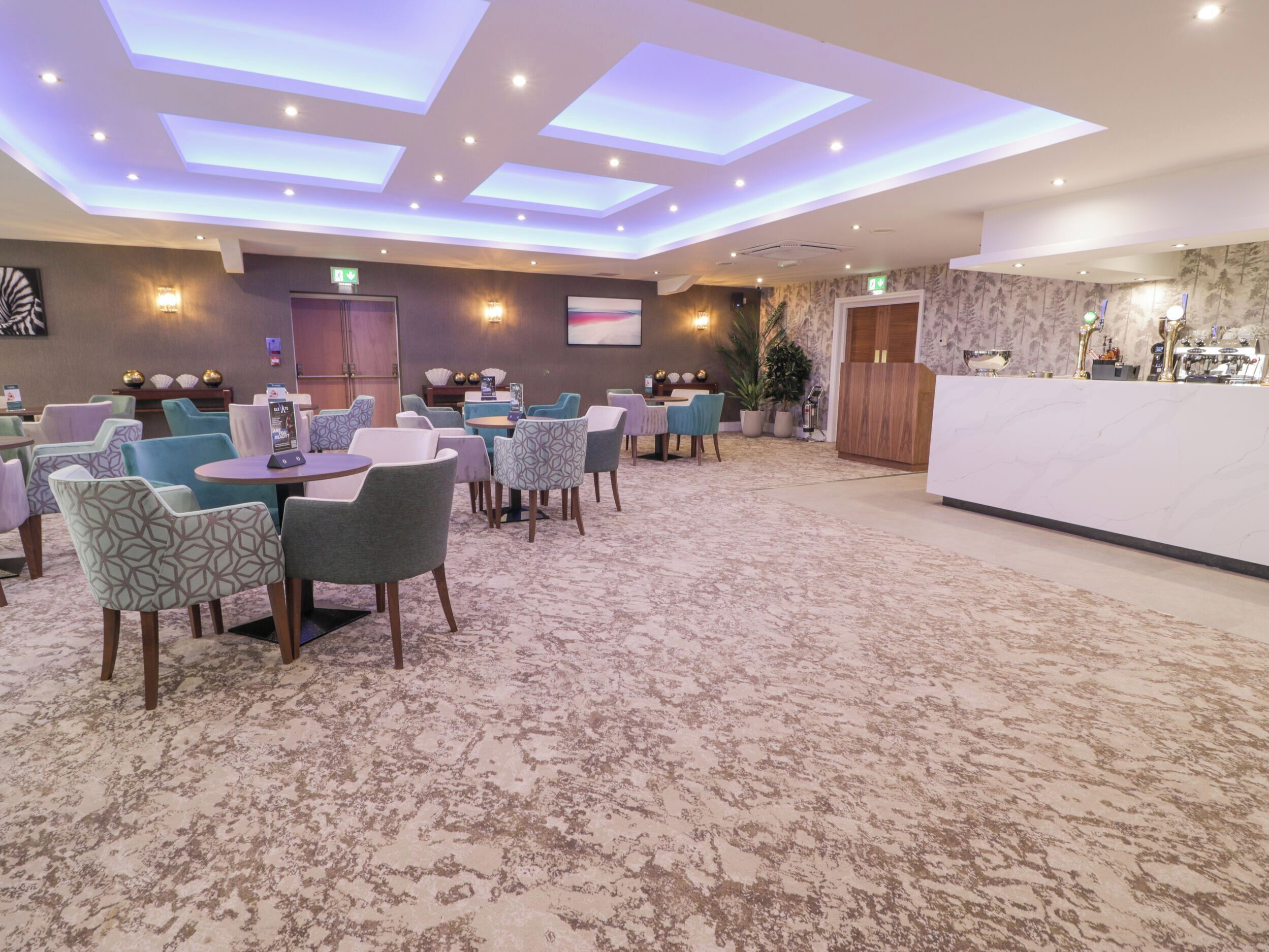 A hotel lobby with a reception desk, multiple seating areas with patterned chairs, and a carpeted floor.