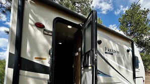 Un modèle Freedom Pilot Express RV, avec une porte ouverte, révélant un petit espace intérieur avec des marches menant à une autre porte, et deux chaises vertes à l’extérieur.