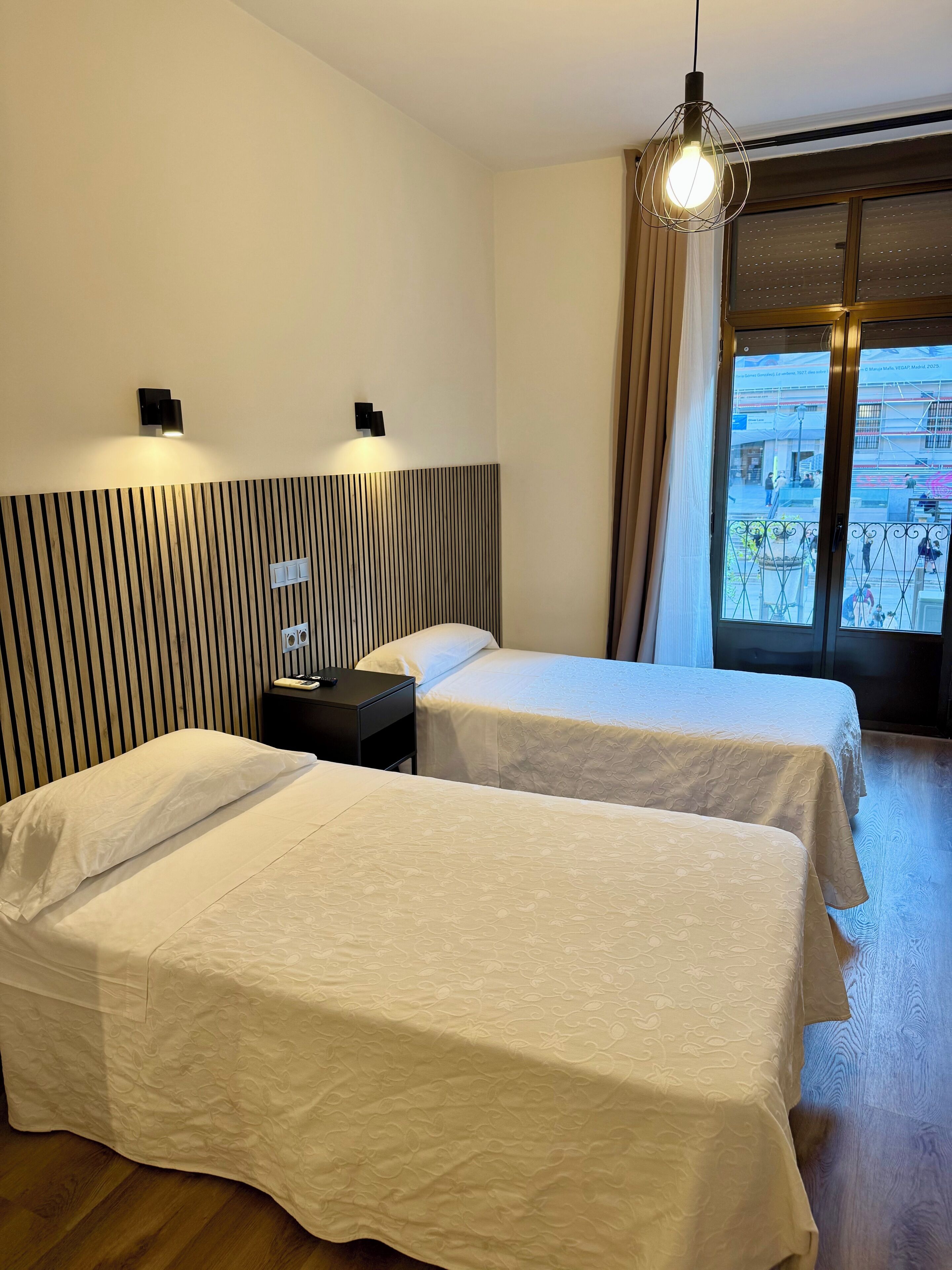 Habitación de hotel con dos camas, cabecero de listones de madera, lámpara colgante y ventana con vista a la ciudad.