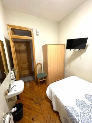 A small, compact room with a wooden floor, a single bed with a patterned bedspread, a wooden wardrobe, a small chair, a wooden door, a sink with a mirror, and a flat-screen TV mounted on the wall. - Hostal Fernández Atocha (Madrid)