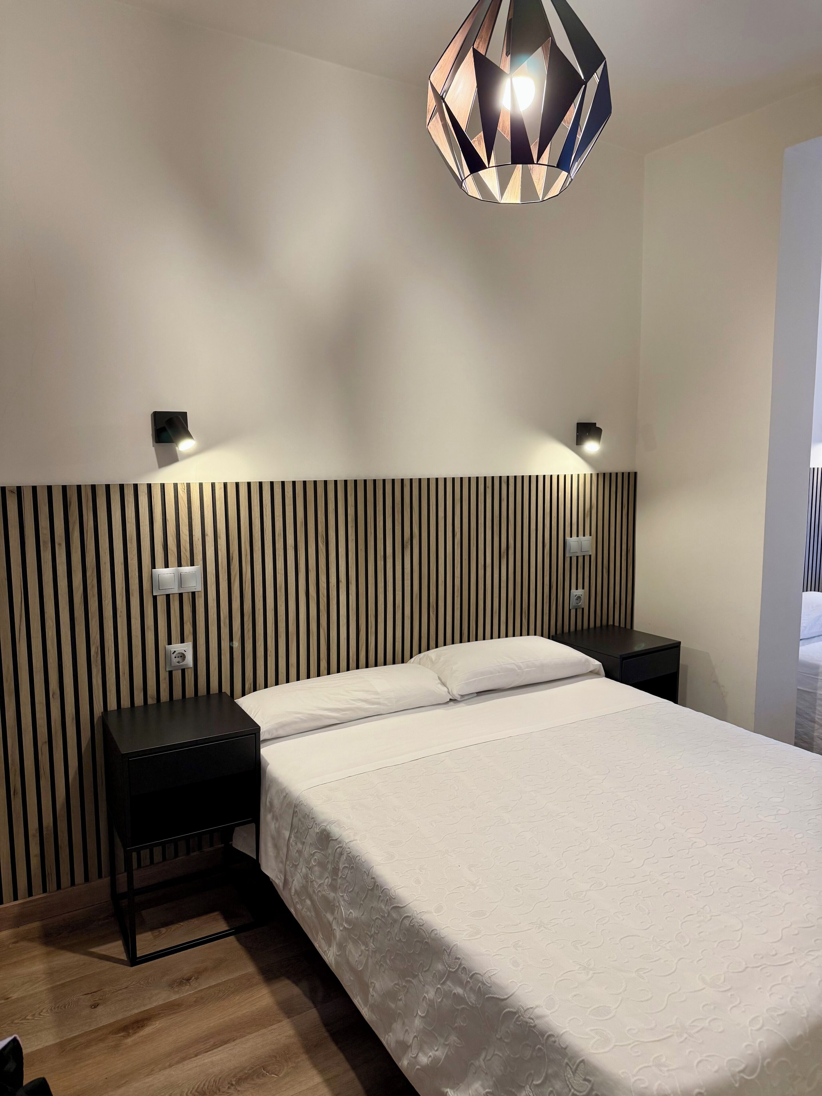 Un dormitorio moderno con una cama grande, dos mesitas de noche, una lámpara de pared y un cabecero de listones de madera.