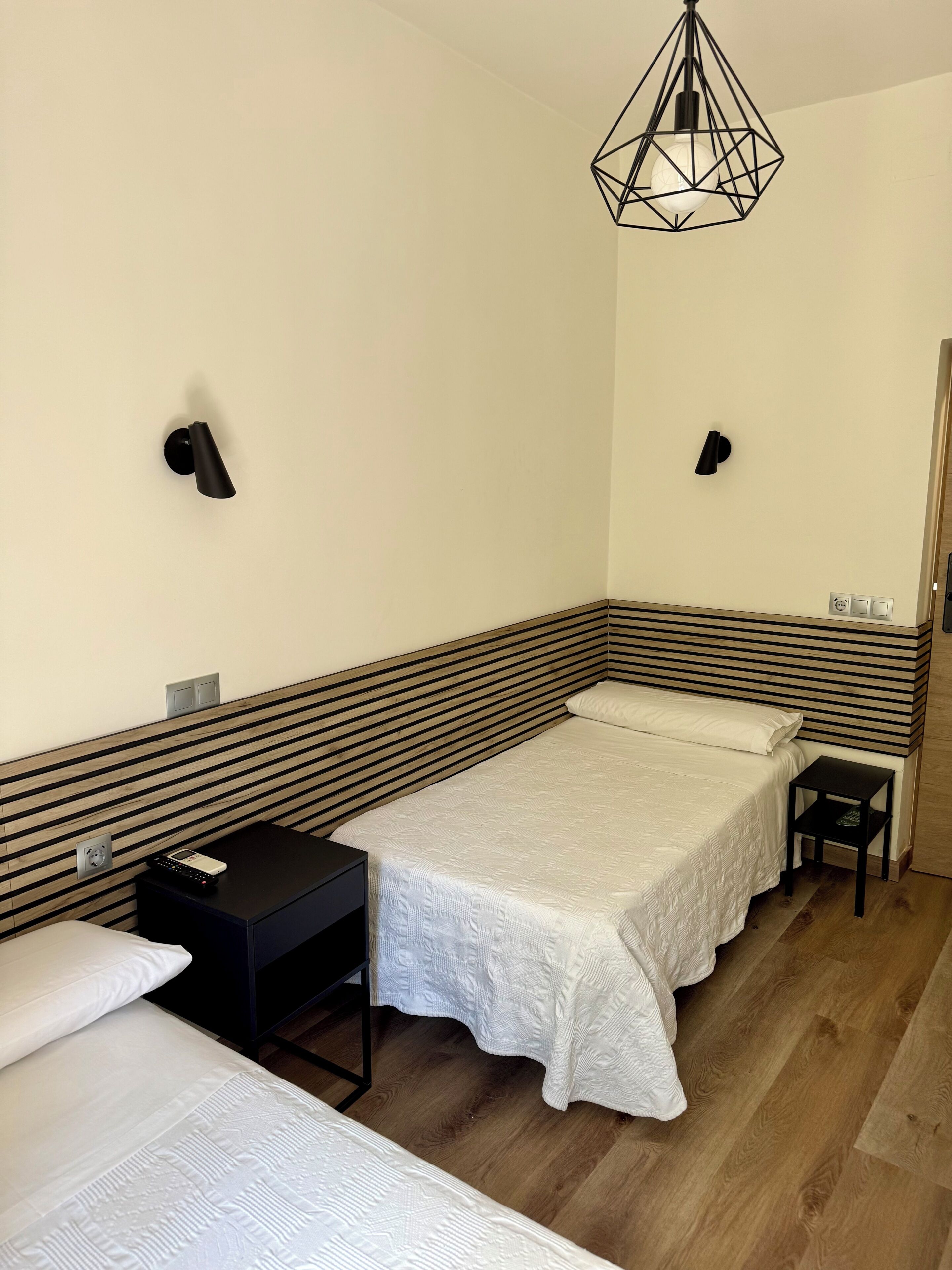 Una habitación de hotel con cama, mesitas de noche, una lámpara de pared y una puerta.