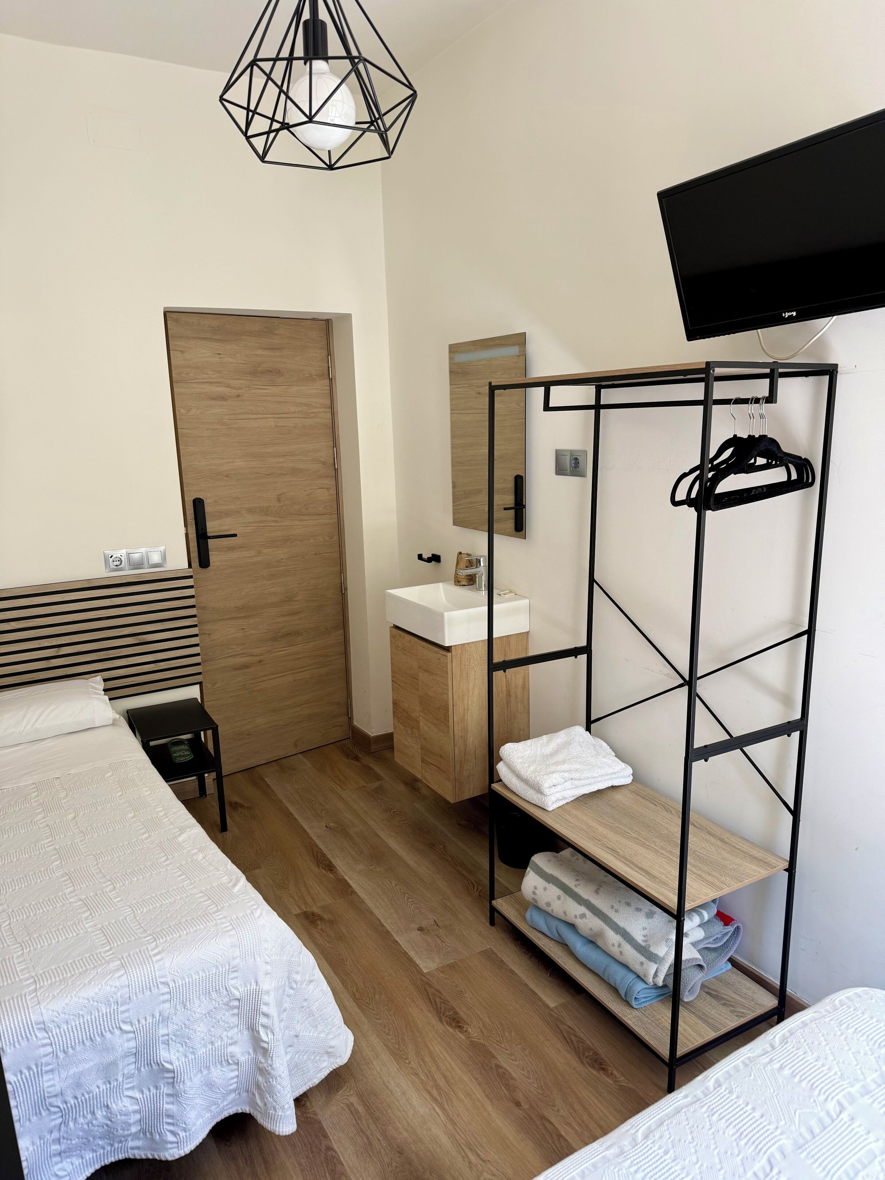 Un dormitorio moderno con una cama, una puerta de madera, un lavamanos blanco, un espejo, un televisor y una estantería metálica.