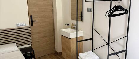 Un dormitorio moderno con una cama, una puerta de madera, un lavamanos blanco, un espejo, un televisor y una estantería metálica.