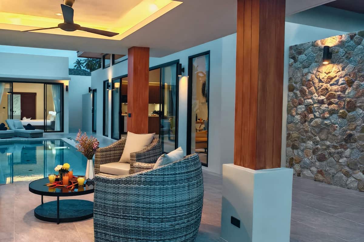 A modern indoor pool area with wicker furniture, a stone wall, and a ceiling fan.