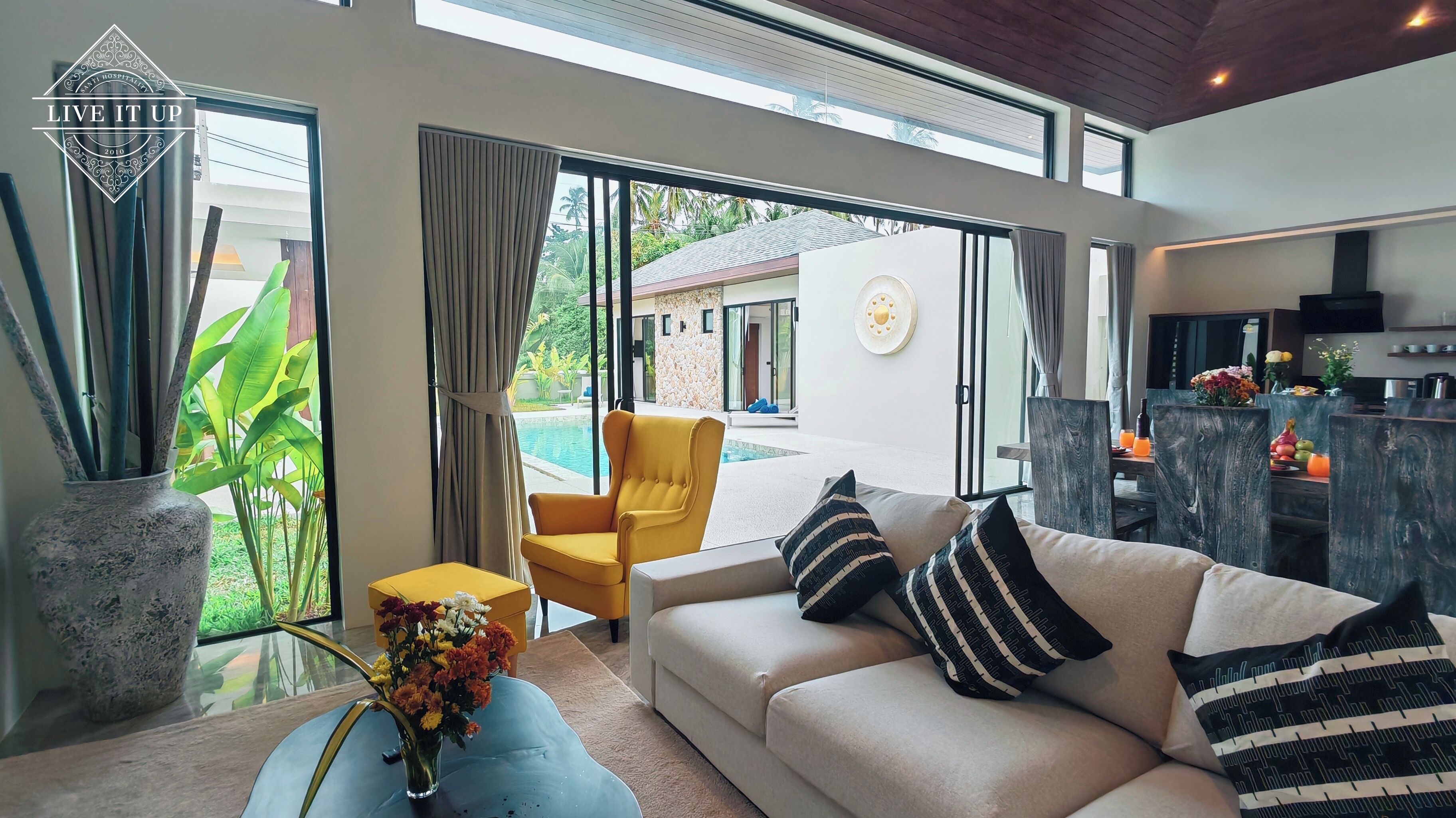 A modern living room with a beige sofa, yellow armchair, and a glass coffee table. There is a large window with curtains, a view of a pool and outdoor area, and a kitchen visible through an open door.