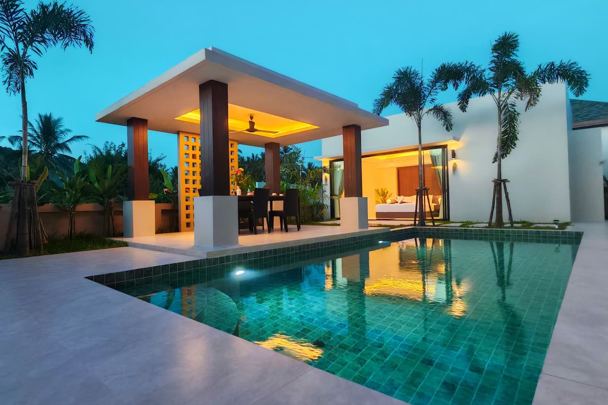 A modern pool area with a covered seating area, a pool with clear blue water, and surrounding tropical plants.