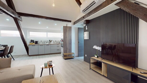 A modern living room with a large flat-screen TV, a wooden coffee table, and a sofa. There is a kitchen in the background with white cabinets and a dining area with a table and chairs.