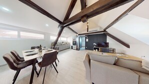A modern loft apartment with exposed wooden beams, a dining area with a table set for four, a kitchenette, a living area with a sofa, and a TV.