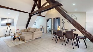 A spacious loft apartment with exposed wooden beams, a large L-shaped sofa, a dining area with chairs, and a view through a glass door to another room.