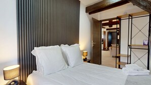 A hotel room with a bed, a nightstand with a lamp, a wooden door, and a view of the hallway.