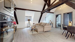 A modern loft-style living space with exposed wooden beams, a large sectional sofa, a dining area, and a kitchenette with a built-in oven.