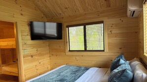A wooden cabin room with a bed, a TV mounted on the wall, and a window with a view of greenery.