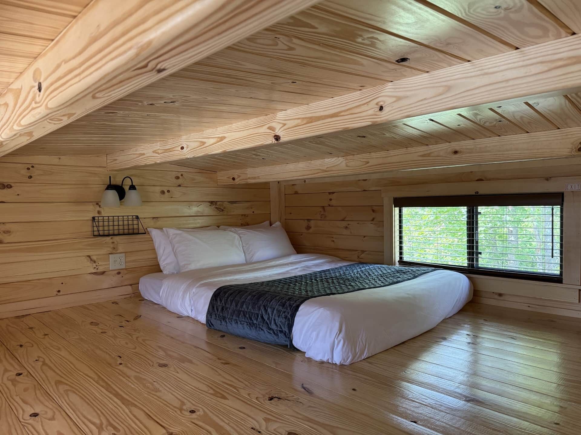 Un dormitorio de cabaña de madera con cama, ventana y lámpara fijada a la pared.