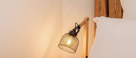 Een netjes opgemaakt bed met witte lakens, een houten hoofdbord en een enkele bedlamp met een metalen kap.