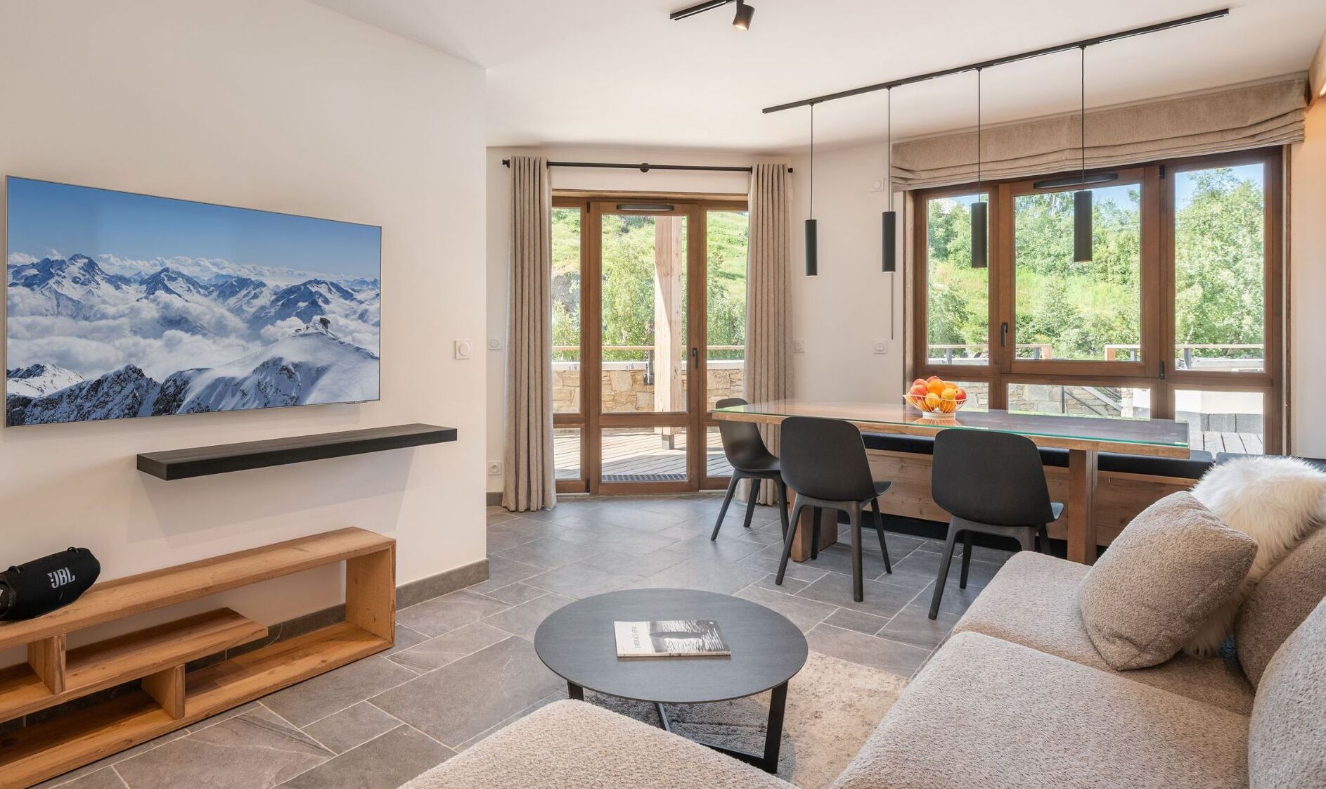A modern living room with a large TV, a wooden bench, a round coffee table, and a sofa. There is a dining area with a table and chairs, and a large window with curtains.