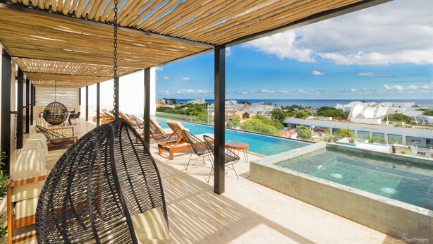 A rooftop pool area with a view of the ocean and cityscape, featuring lounge chairs and a hanging chair.