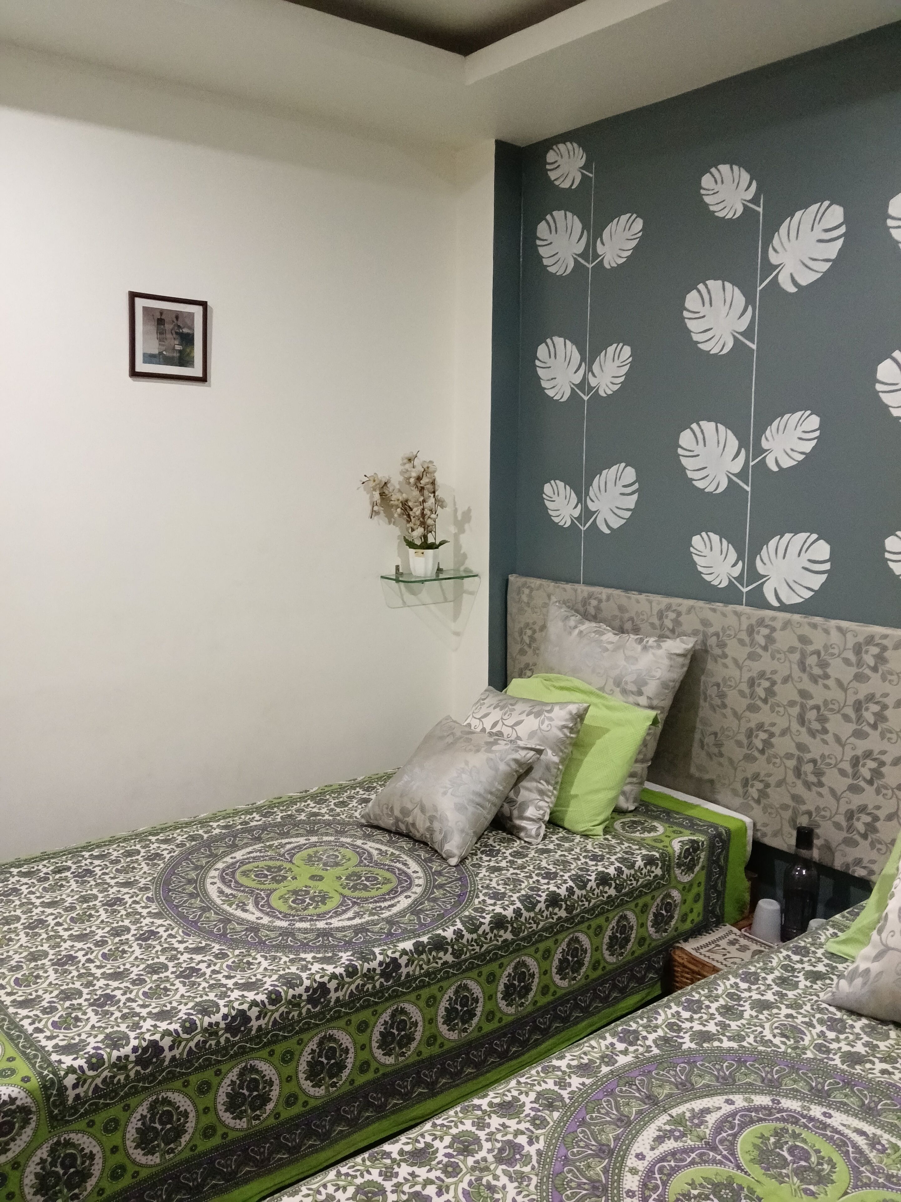 A bedroom with a patterned bedspread, a headboard with a leaf design, a small shelf with a vase, and a framed picture on the wall.