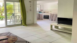 Amplia zona de estar con un puertas correderas de vidrio que dan a un balcón, una cocina con cortinas estampadas y un televisor de pantalla plana sobre un mueble.