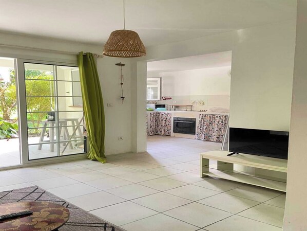 A spacious living area with a sliding glass door leading to a balcony, a kitchen with patterned curtains, and a flat-screen TV on a stand.