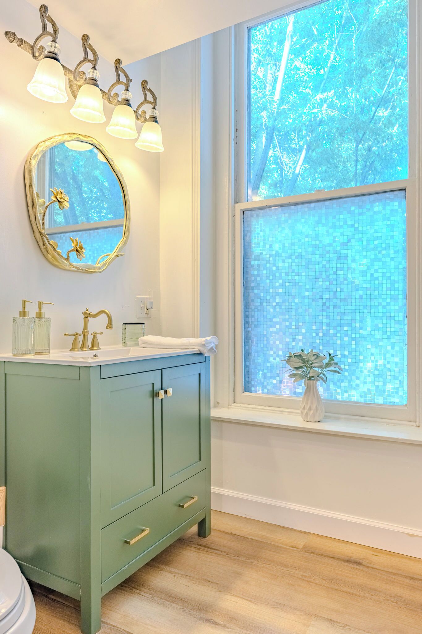 Une salle de bain avec un meuble-lavabo vert, un miroir rond avec un cadre doré, un lavabo blanc et une fenêtre donnant sur les arbres.