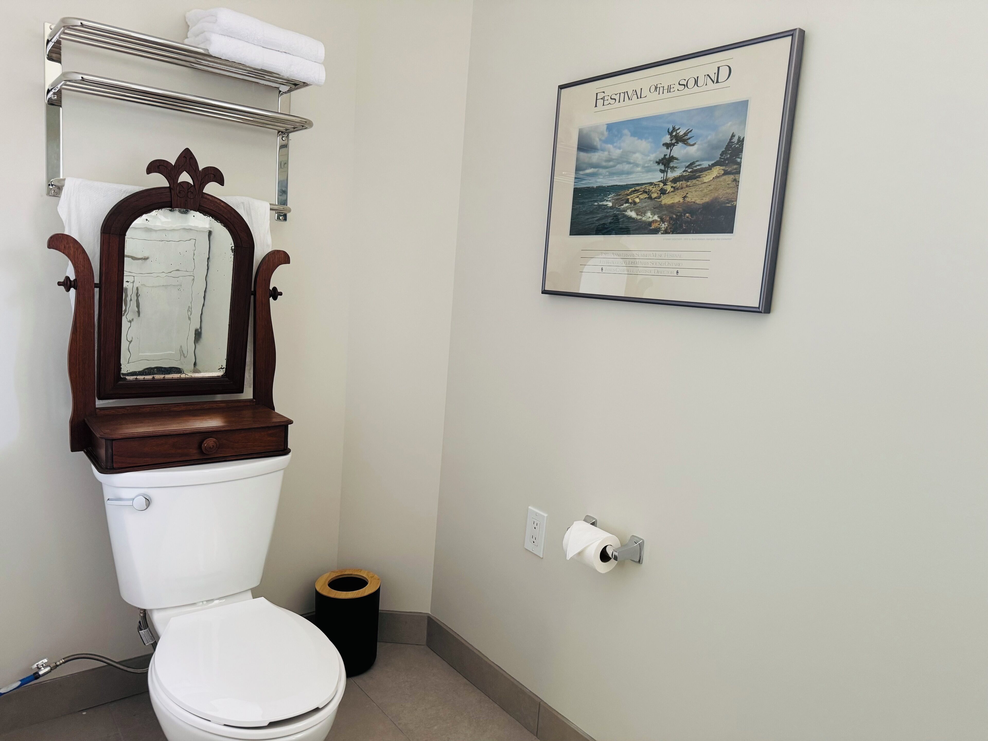 A bathroom with a white toilet, a wooden-framed mirror, a towel rack with a towel, a framed picture on the wall, and a toilet paper holder.